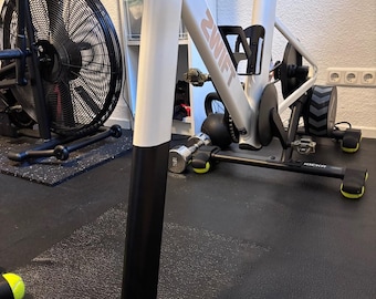 May include: A white and black stationary exercise bike with the word "ZWIFT" printed on the frame. The bike has a water bottle holder and a black fan in the background. The bike is on a black mat.