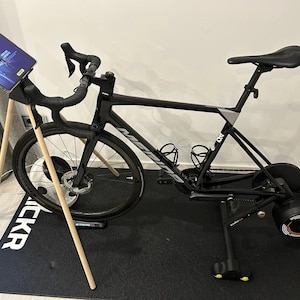 Könnte beinhalten: Ein schwarzes Rennrad mit einem Tablet am Lenker ist an einem schwarzen Indoor-Fahrradtrainer mit einem roten und orangen Rad befestigt. Das Fahrrad steht auf einer schwarzen Matte mit dem Wort "WACKR" darauf.
