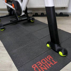 以下が含まれることがあります： 赤い文字で「RIDE THINK」と書かれた黒いエクササイズマット。マットの下には、黒と黄色のアクセントが付いた固定式エクササイズバイクがあります。バイクは白いフレームと黒いコンポーネントを備えています。マットはインターロッキングタイルでできています。