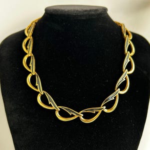 Può includere: Una collana dorata con dettagli in smalto nero. La collana presenta una serie di maglie a forma di goccia, interconnesse. Il gioiello è esposto su un busto di manichino nero, che ne mette in risalto il design e lo stile.