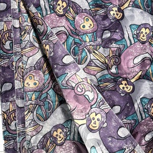 May include: Fabric with a repeating pattern of cartoon monkeys in shades of purple, gray, teal, and yellow. The monkeys have smiling faces and are surrounded by stylized leaves and vines. The fabric is folded, showing the pattern's repeat.