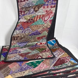 May include: A patchwork table runner with a black border. The runner is made of various fabric scraps in a variety of colors and patterns, including red, blue, green, and purple. The runner is rectangular and appears to be handmade.