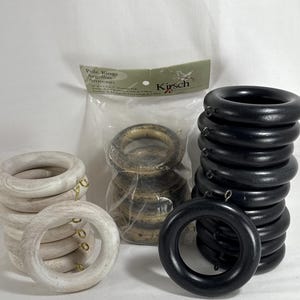 May include: A collection of wooden curtain rings in various colors. There are stacks of black, white, and natural-toned rings. Some rings have small gold-colored metal hooks. A package of rings is visible with the text "Pole Rings".