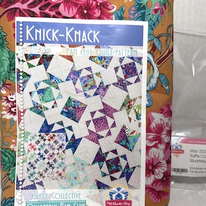 May include: A Knick-Knack Fan Club quilt pattern document featuring a geometric design with white polka-dot fabric and colorful floral prints, placed over a folded piece of mustard yellow fabric with a large pink and green floral pattern.