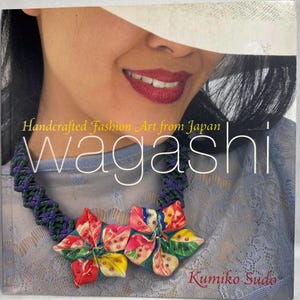 May include: A book cover titled Handcrafted Fashion Art from Japan wagashi by Kumiko Sudo. The cover features a person wearing a necklace with a dark purple and green braided cord and two large, multi-colored fabric flower-shaped pendants.
