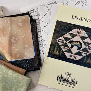 Peut inclure: Un ensemble de morceaux de tissu de différentes couleurs et motifs, dont du bleu, du marron et du beige moucheté. Un livret de patrons intitulé "LEGENDS" avec un motif d'ours et de forêt accompagne le tissu. Le livret est de "Granola Girl".