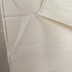 May include: Off-white fabric with subtle horizontal lines and a textured appearance. The fabric has a raw edge and a gray border on one side. The material is folded over, revealing more of the textile.