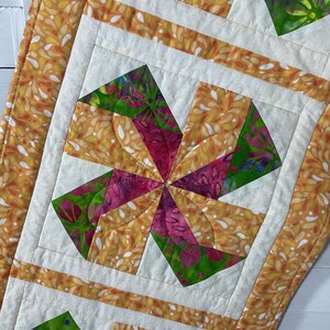 May include: A quilted table runner featuring a central pinwheel block with vibrant green, magenta, and orange batik fabric. The border consists of cream fabric with an orange teardrop pattern, accented by straight-line machine quilting throughout.
