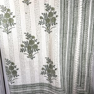 May include: A white cotton quilt featuring a repeating pattern of sage green floral motifs and vertical running stitch lines. The quilt has a wide border with a geometric sage green pattern and a blue plastic clip attached to the top edge.