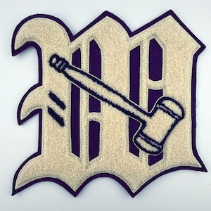 May include: A felt patch featuring a large, cream-colored letter M in an old English font with a dark purple border. Overlaid on the letter is a dark purple embroidered gavel icon, positioned diagonally across the center of the letter.
