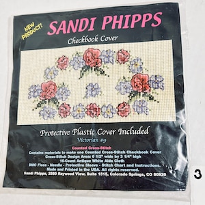 May include: A Sandi Phipps cross-stitch checkbook cover kit with a floral design of red and purple flowers. Includes a protective plastic cover. The cross-stitch area is 16.5 cm wide by 8.3 cm high. Made in the USA. The kit is for a Victorian #9 design.