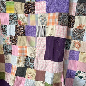 May include: A patchwork quilt featuring a variety of fabric squares in shades of purple, pink, orange, green, and brown with floral, plaid, and polka dot patterns. A large, dark purple letter B is appliqued onto the center of the quilt surface.