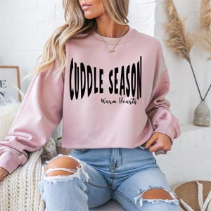 May include: A light pink sweatshirt with the words "Cuddle Season" in a black, wavy font. Below the text is "Warm Hearts" in a smaller, cursive font. The sweatshirt has long sleeves and a crew neck.