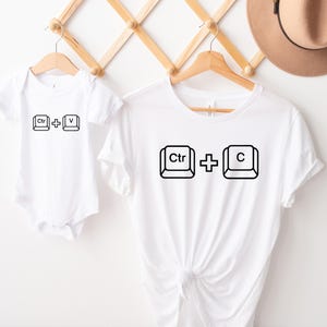 May include: White t-shirts and a baby onesie with black keyboard shortcut graphics. The adult t-shirt features "Ctrl + C", while the onesie displays "Ctrl + V". A brown hat hangs in the background.