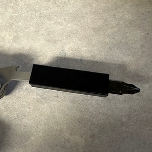 Puede incluir: Una multiherramienta plateada con un accesorio rectangular negro y una punta de destornillador Phillips. La herramienta también tiene un abridor de botellas. La imagen es un primer plano.