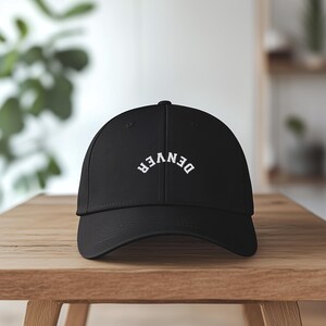 May include: Black baseball cap with the word "DENVER" arched across the front in white lettering. The cap is sitting on a wooden surface. The background is blurred, with hints of a plant and shelves.
