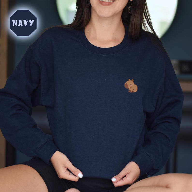 May include: A navy blue sweatshirt with a small brown capybara embroidered on the left chest. The sweatshirt has a crew neck and long sleeves.