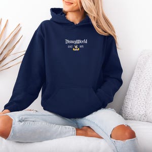 May include: Navy blue hooded sweatshirt with the text "DisneyWorld" and "EST. 1971" in white, along with a small Mickey Mouse graphic. The sweatshirt has a front pocket and a drawstring hood. The model is wearing light blue jeans.