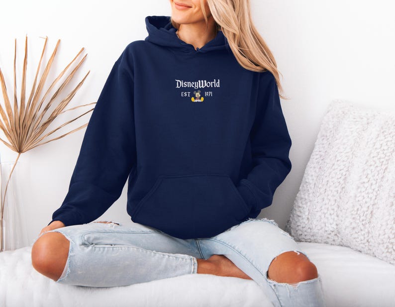 May include: Navy blue hooded sweatshirt with the text "DisneyWorld" and "EST. 1971" in white, along with a small Mickey Mouse graphic. The sweatshirt has a front pocket and a drawstring hood. The model is wearing light blue jeans.