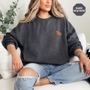May include: A dark heather grey sweatshirt with a small embroidered capybara on the left chest. The sweatshirt is being worn by a person sitting on a white couch.
