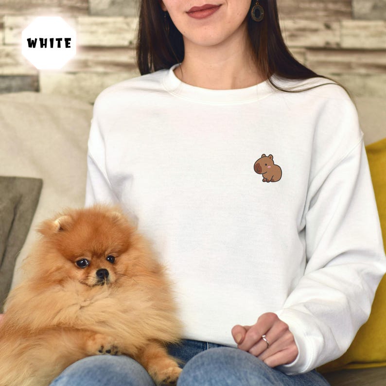 May include: White sweatshirt with a small brown capybara illustration on the left chest. The sweatshirt is being worn by a person with long brown hair and a small dog sitting on their lap.