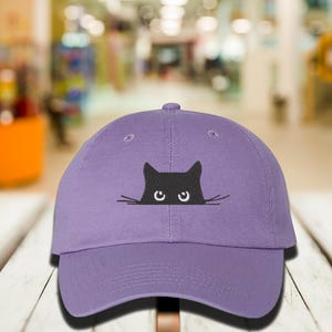 May include: A purple baseball cap with a black cat peeking over the brim. The cat has white eyes and whiskers.