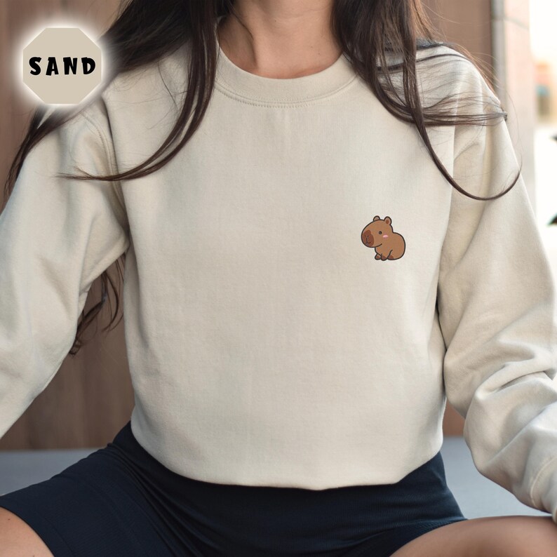 May include: A beige sweatshirt with a small brown capybara graphic on the front. The sweatshirt has a crew neck and long sleeves.