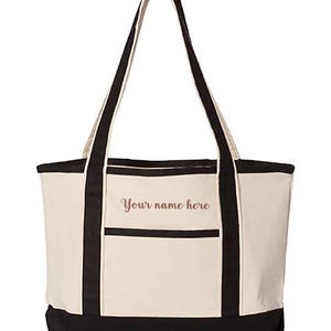 May include: Cream-colored tote bag with black accents and handles. Features a front pocket and the text "Your name here" in a script font. The bag is designed for carrying items.