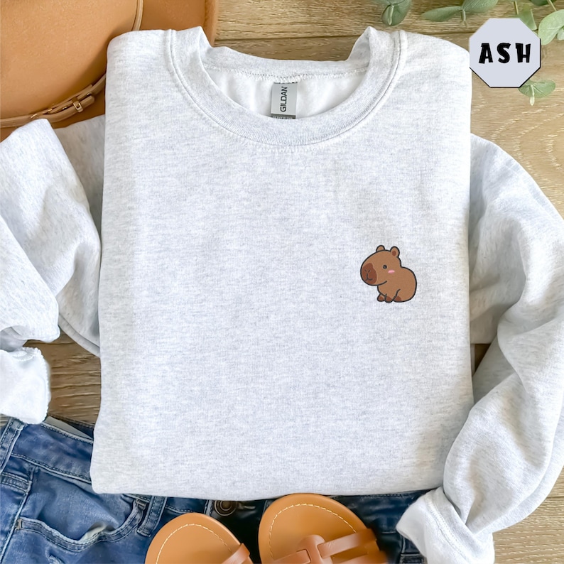 May include: A gray sweatshirt with a small brown capybara illustration on the left chest. The sweatshirt is laid flat on a wooden surface with a pair of brown sandals and blue jeans.