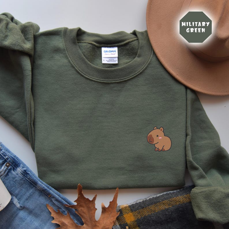 May include: A green sweatshirt with a small brown capybara graphic on the left side. The sweatshirt is laid flat on a white surface with a pair of blue jeans and a brown hat in the background.