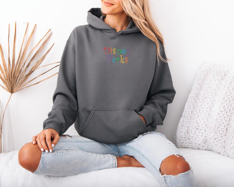 Puede incluir: Una sudadera con capucha gris antracita con un bolsillo delantero y una capucha. La sudadera tiene el texto bordado "Disney Parks" en colores arco&iacute;ris. La modelo lleva vaqueros azul claro con rodillas rasgadas.