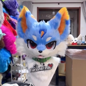 May include: A blue, white, and orange furry fox head with large red eyes, a green collar, and the text "Fursona Finds." The head is mounted on a white form. The fox head has a blue and orange design on the ears.