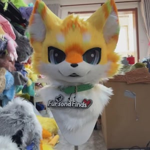 Puede incluir: Una cabeza de fursuit de zorro amarillo, naranja y blanco con grandes ojos azules y un collar verde. El fursuit tiene el texto "Fursona Finds" en el pecho. Las orejas tienen detalles verdes y amarillos.