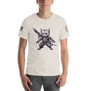 May include: A white t-shirt with a cartoon image of a cat wearing military gear and holding a rifle. The cat is wearing a helmet, body armor, and tactical gear. The cat is standing in a fighting stance. The t-shirt has a small American flag on the left sleeve and a small patch on the right sleeve.