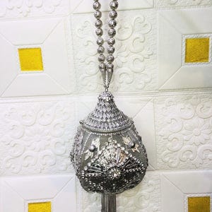 May include: A silver-toned, beaded handbag with a beaded handle and a tassel. The bag is embellished with rhinestones and decorative metalwork. The dimensions are 12 cm x 7 cm x 14 cm. The bag is hanging against a white textured wall with yellow accents.