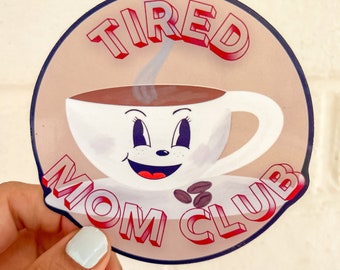 Puede incluir: Una pegatina redonda con una taza de café de dibujos animados con una cara sonriente y el texto "TIRED MOM CLUB" en letras rojas y blancas.