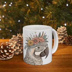 May include: White ceramic mug with a gray and white macaw wearing a floral crown of pink, yellow, and white flowers. The mug has a white handle.