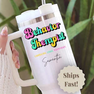 May include: White tumbler with a handle, featuring the words "Behavior Therapist" in colorful, retro-style lettering. Below the text is "Changing Lives, Everyday" and a name in script. The tumbler has a silver band.