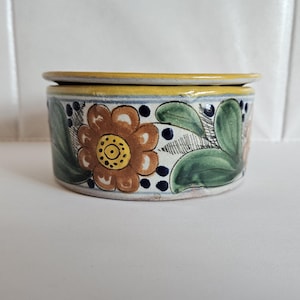 May include: A round ceramic butter dish with a lid, featuring a hand-painted floral pattern in shades of brown, green, and yellow, accented with small dark blue dots along the rim and a solid yellow band around the top edge of the lid.