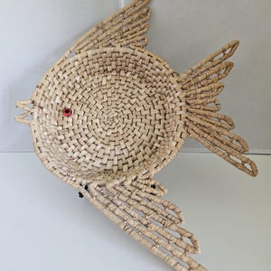 May include: A woven fish-shaped basket with a round body and extended fins. The basket is made of natural-colored woven material with a red bead eye. The fish is designed to be a decorative item.