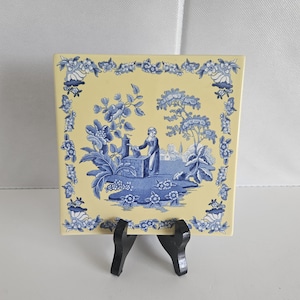 May include: A square ceramic tile with a pale yellow background and a blue transferware-style illustration. The design features a central scene of a figure at a well surrounded by trees and flowers, framed by a decorative floral border pattern.