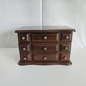 May include: A dark brown wooden jewelry box featuring three horizontal drawers. Each drawer is accented with a small, round, silver-toned metal knob with a floral pattern. The box has a smooth, polished finish and a rectangular shape.