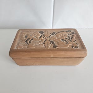 May include: A rectangular terracotta box with a lid featuring a carved relief design of two symmetrical figures facing each other. The natural clay material has a light brown tone with dark brown accents highlighting the intricate patterns.