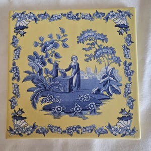 May include: Square ceramic tile with a yellow background and a blue and white pastoral scene. The scene depicts a woman at a well, surrounded by floral and foliate elements. The border features a floral design.