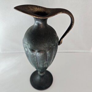 May include: A small, antique-style metal pitcher with a dark, aged finish. The pitcher has a wide, flared spout and a curved handle. It stands on a round base. The metal has a mottled appearance, suggesting age and use. The pitcher is likely decorative.