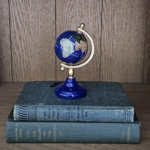 May include: A small decorative globe featuring a deep blue ocean and inlaid stone continents in shades of tan, green, and white. The globe is mounted on a gold-toned metal meridian arc and a circular blue base, resting atop two stacked hardcover books.