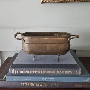 May include: An oval-shaped, metallic bronze-toned planter with a smooth finish, featuring two curved handles on the sides and four small, sturdy legs. The vessel has a deep, open basin and a slightly weathered, aged appearance throughout its surface.