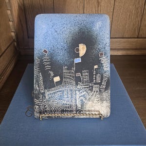 May include: A rectangular ceramic plaque featuring a speckled blue and black gradient background with white etched line drawings of buildings and structures. The design includes small gold and blue geometric accents and flags atop the structures.