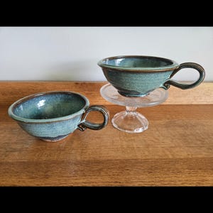 May include: Two handmade ceramic teacups with a teal glaze and brown trim. One cup sits on a clear glass pedestal. The cups have a rounded shape with a handle. The background is a wooden surface and a white wall.