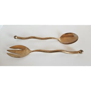 May include: A pair of gold-toned metal salad serving utensils consisting of one spoon and one three-pronged fork. Both utensils feature a long, slender, wavy handle that terminates in a rounded, spherical knob at the end of each handle.